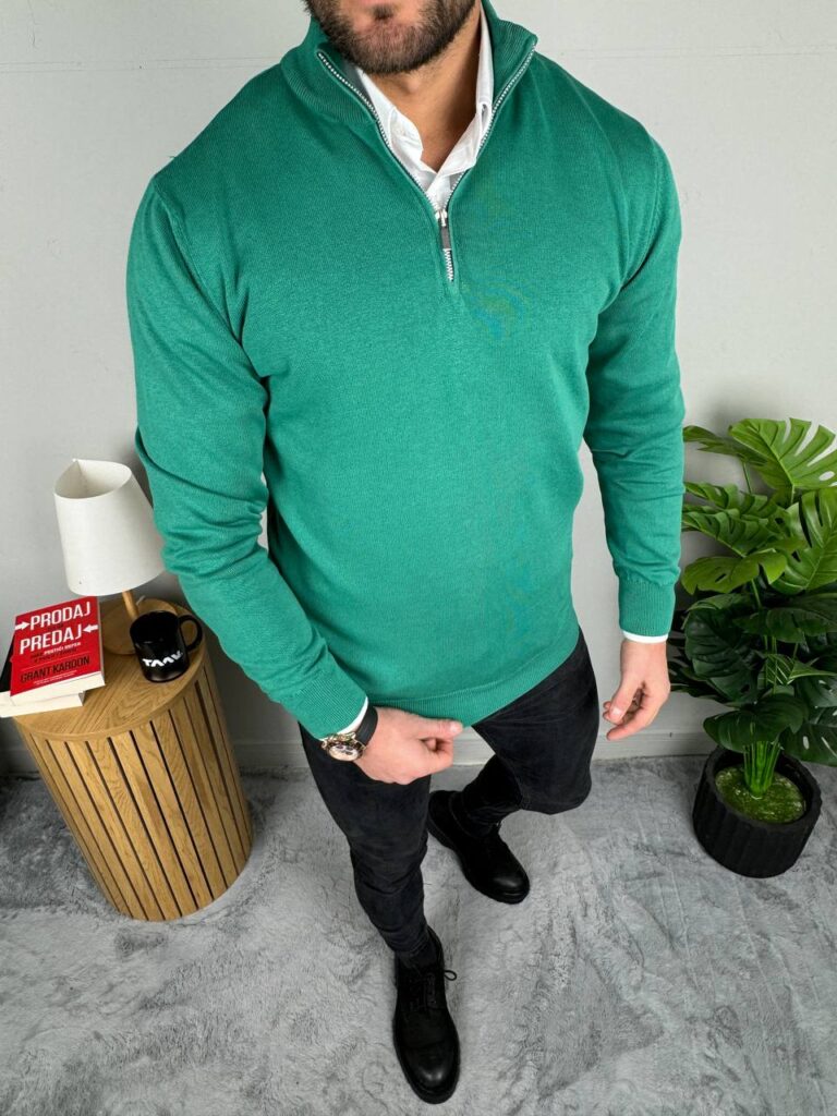 Muški džemper polu zip u zelenoj boji uz crne pantalone, ceo lik ispred dekoracije sa biljkom