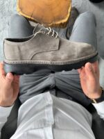 Muške cipele sive boje od brušene kože koje model drži iznad zelenih pantalona